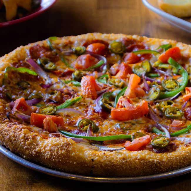 A restaurant-style pizza with tomatoes, green peppers, jalapeños, and red onions on a metal tray.