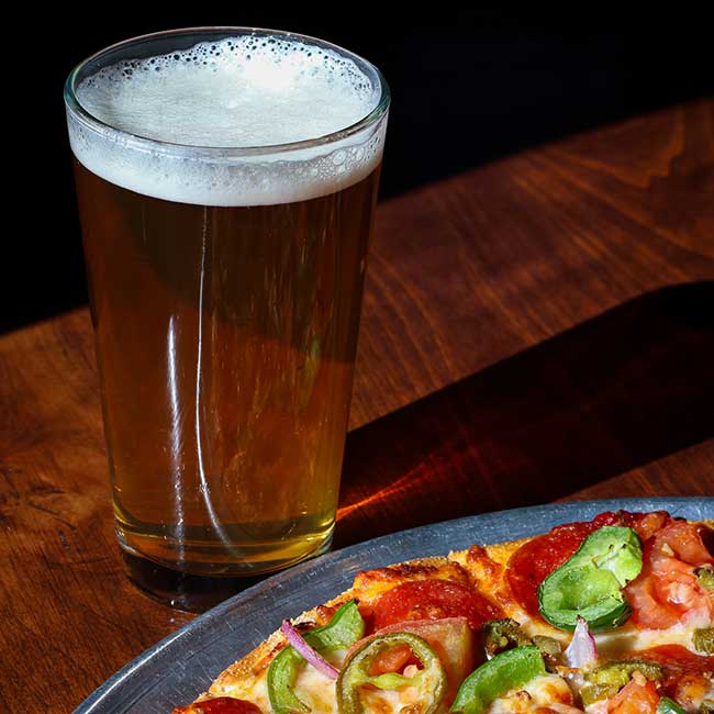 A glass of beer in Boise, Idaho, next to a plate of pizza with peppers on a wooden table.
