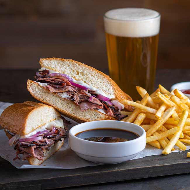 French dip sandwich with fries, au jus, and beer in Boise, Idaho on a wooden table.